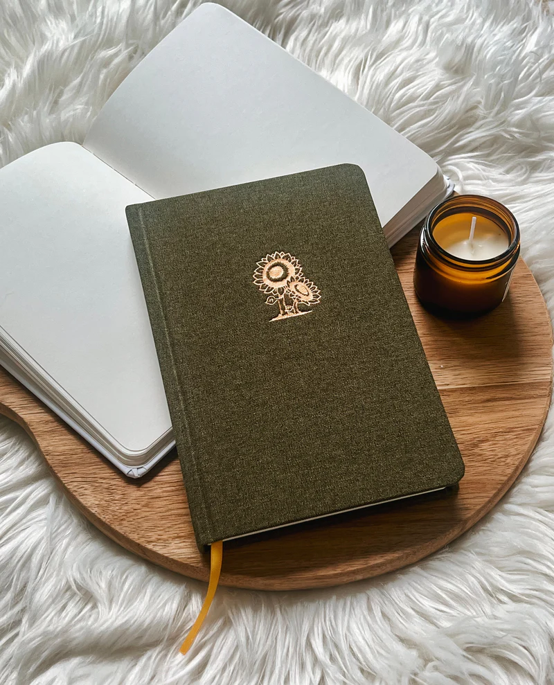 Fabric hardcover journal with gold sunflower design placed on wooden tray with candle and open notebook.