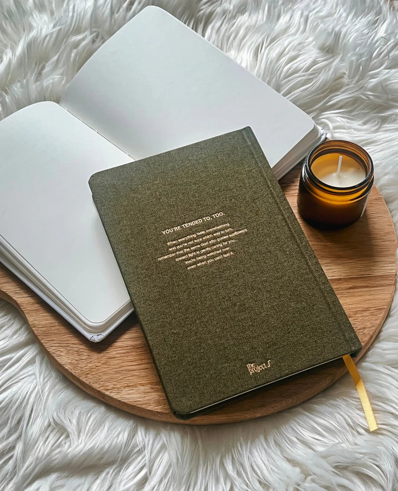 Olive green fabric journal with gold text on back cover by Project J placed on wooden tray beside open notebook and candle.