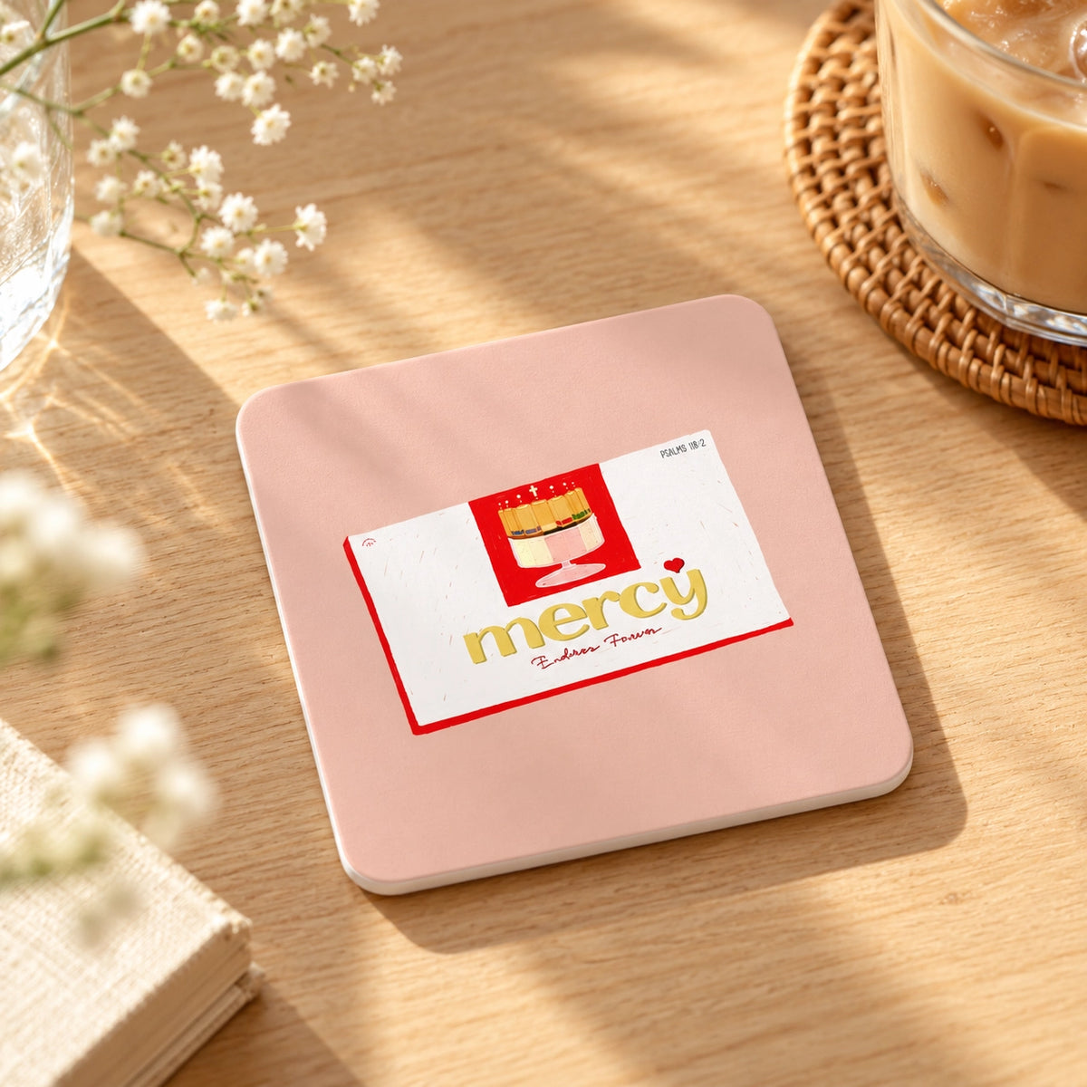 Pink square Christian coaster with mercy artwork styled on a sunlit wooden table beside iced coffee and flowers.