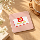 Pink square Christian coaster with mercy artwork styled on a sunlit wooden table beside iced coffee and flowers.
