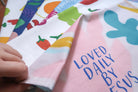 Pastel pink tea towel with the blue phrase "LOVED DAILY BY JESUS" and colorful shapes, a Christian home decor gift from The Commandment Co.