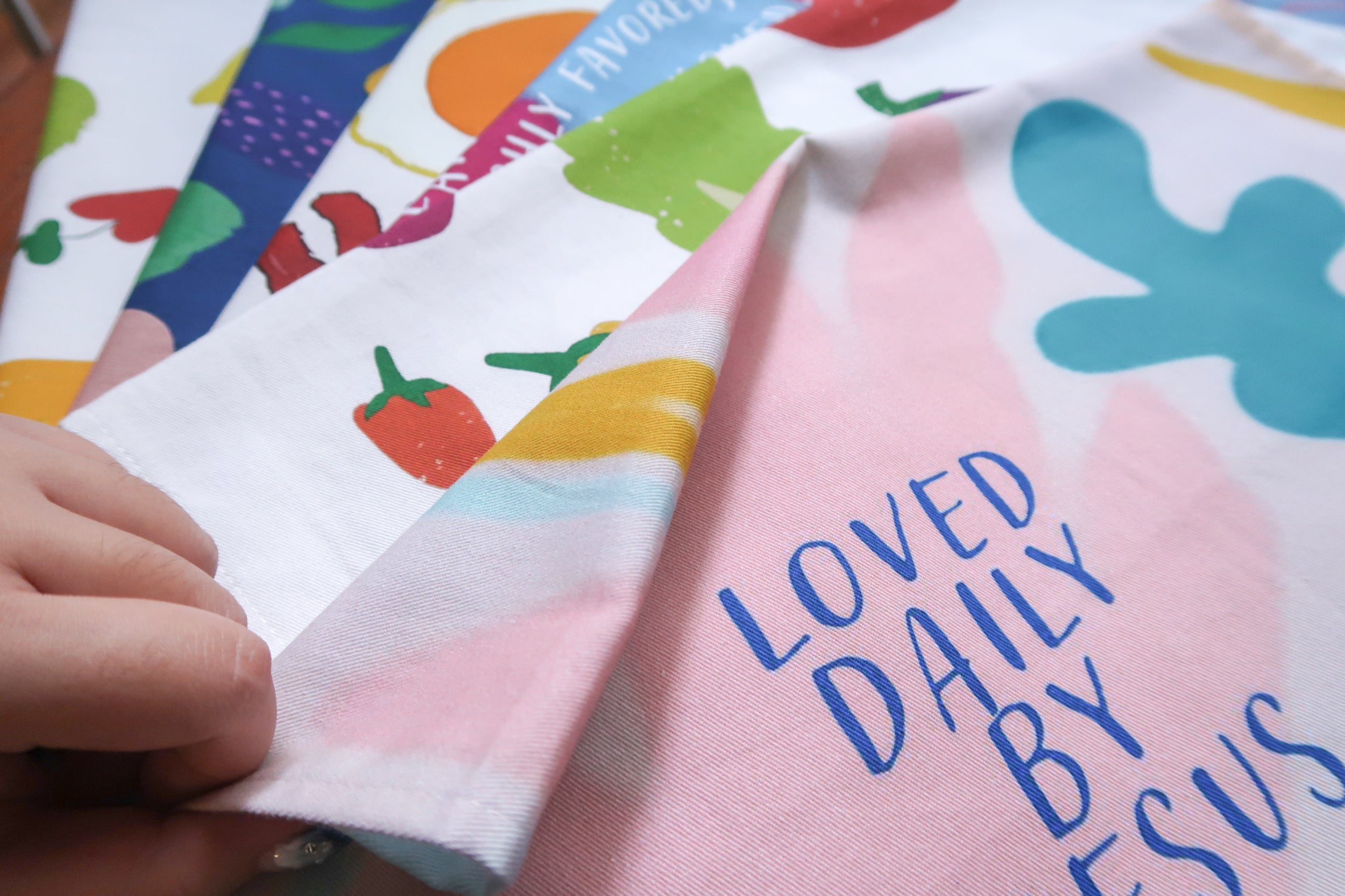 Pastel pink tea towel with the blue phrase "LOVED DAILY BY JESUS" and colorful shapes, a Christian home decor gift from The Commandment Co.