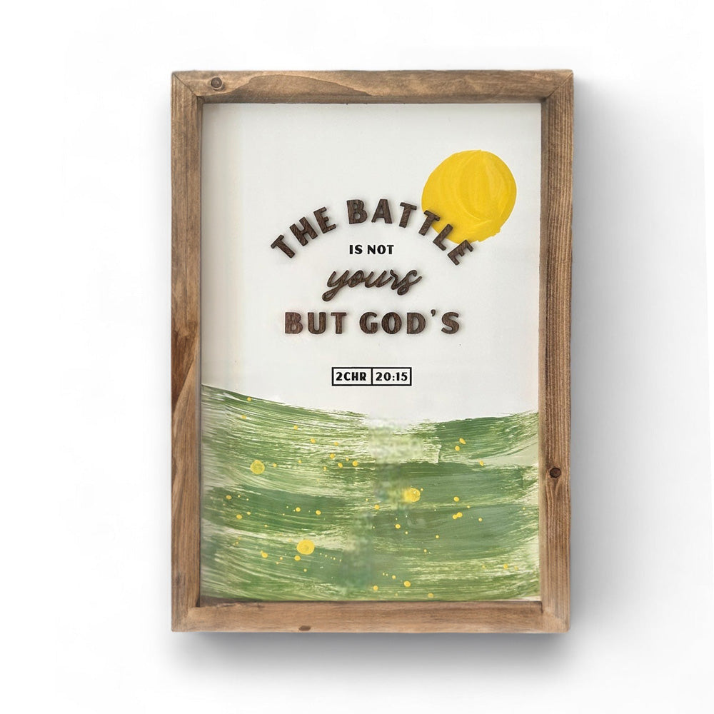 A framed Christian poster with the verse 'The battle is not yours but God's' from 2 Chronicles 20:15. The design features a stylized sun and a green abstract landscape, symbolizing faith and encouragement. The wooden frame adds a rustic, natural feel.