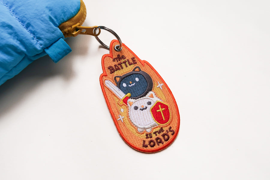 Embroidered Christian warrior cat keychain with “The Battle Is The Lord’s” text, featuring sword and cross shield on orange background.