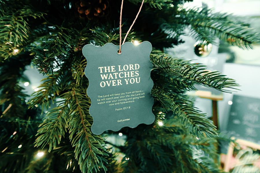 Christian Christmas tree hanging ornament featuring the Bible verse 'The Lord Watches Over You' from Psalm 121:7-8, with a green scalloped design and twine string on a decorated tree.