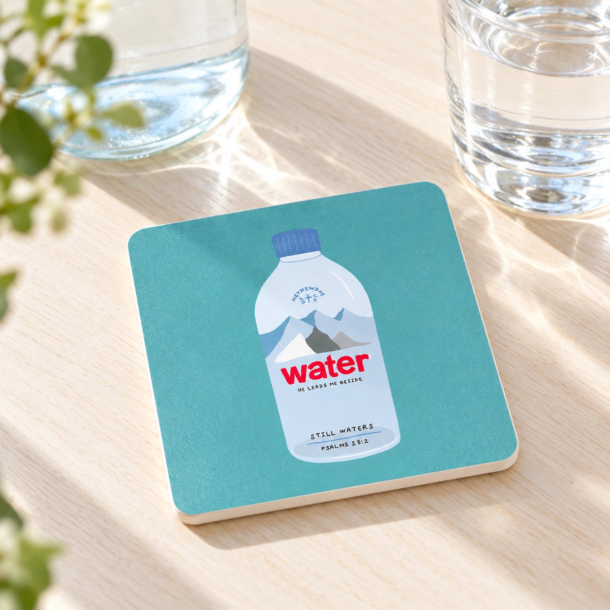 The Still Waters Psalm 23:2 Christian coaster styled on a light wooden table beside glasses of water.