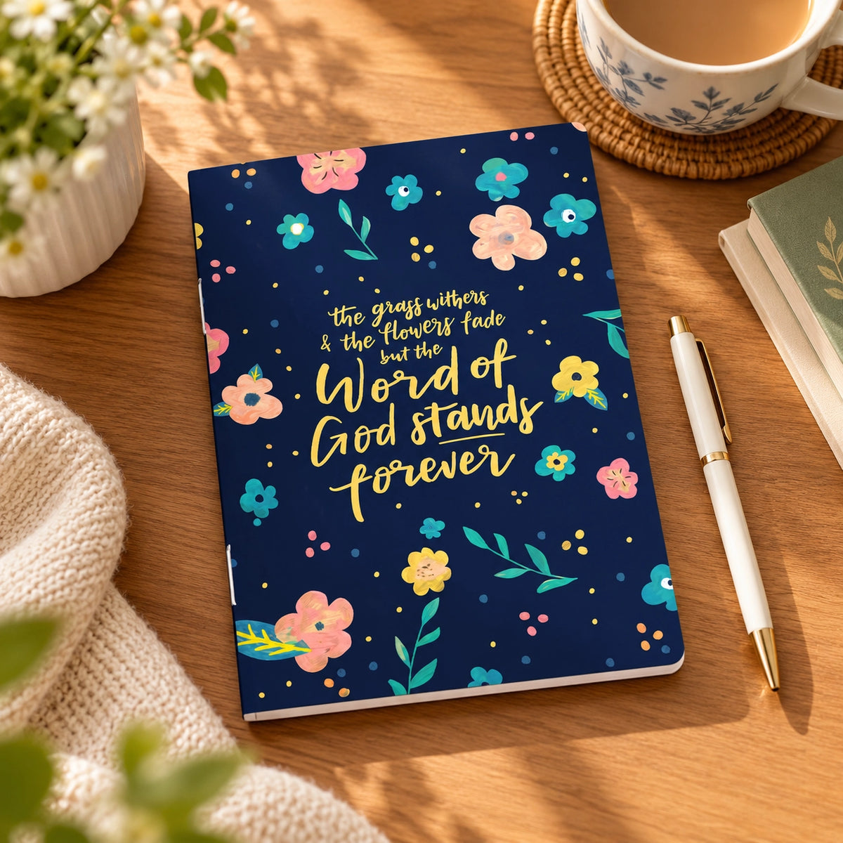 The Word of God Stands Forever A6 Christian notebook styled on a warm wooden desk with flowers, tea and a pen.