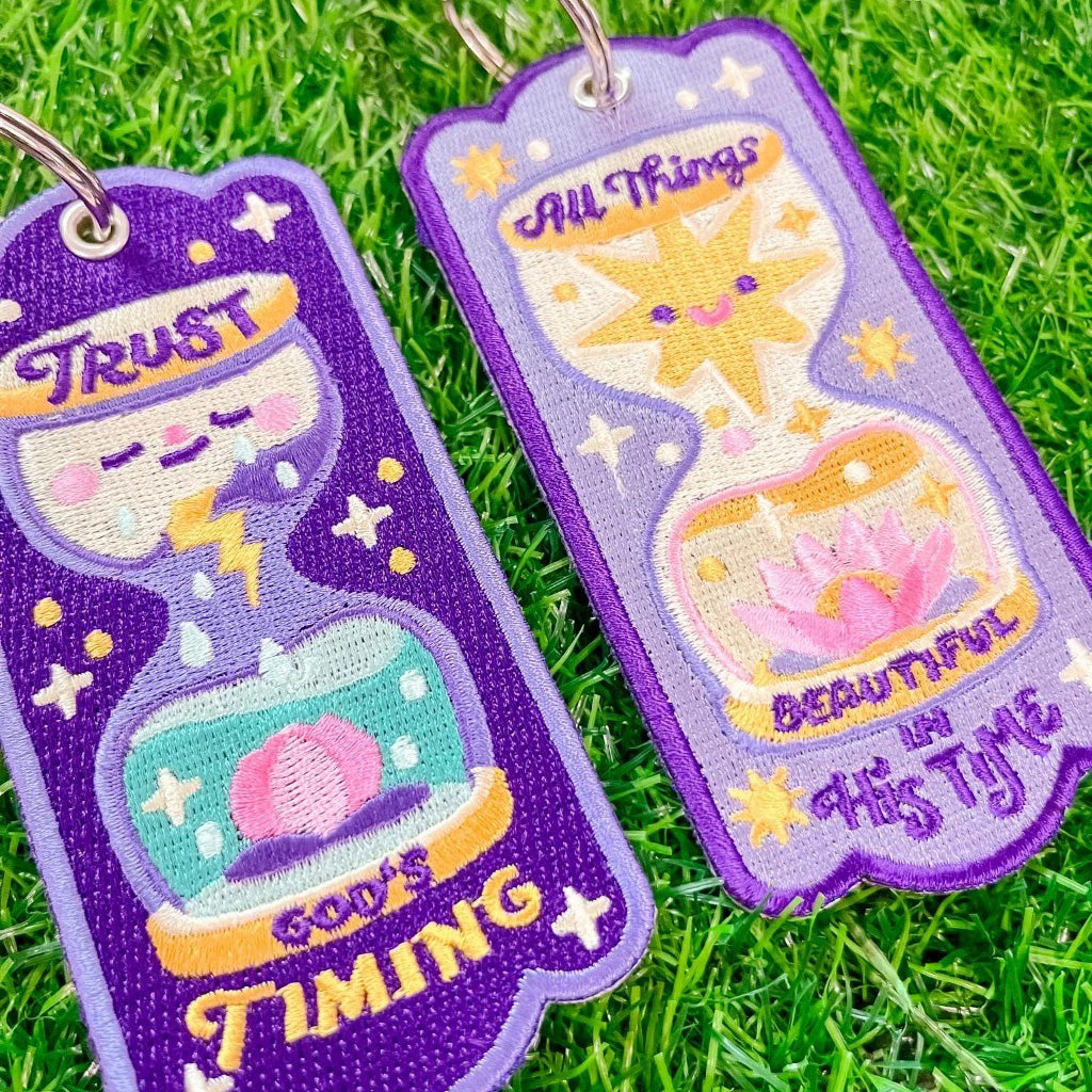 Two embroidered hourglass-shaped keychains with Bible messages on green grass. One says 'Trust God's Timing' with a storm and blooming flower design, and the other says 'All Things Beautiful in His Time' featuring a bright sun and lotus flower illustration.