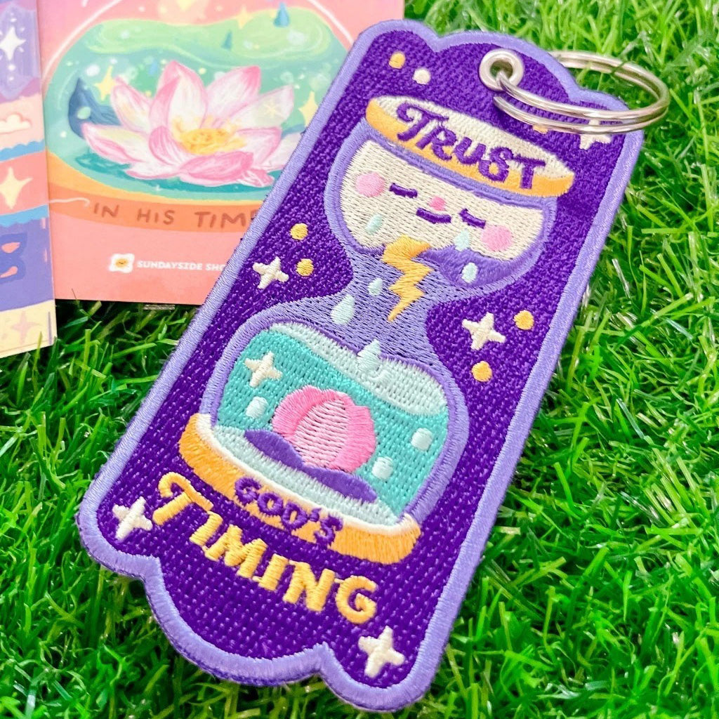 Purple embroidered keychain featuring an hourglass design with the message 'Trust God's Timing,' a blooming flower, raindrops, and lightning, placed on green grass with a card in the background.