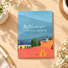 By His Stripes We Are Healed Isaiah 53:5 Christian card lying flat on a warm wooden desk with flowers and pen