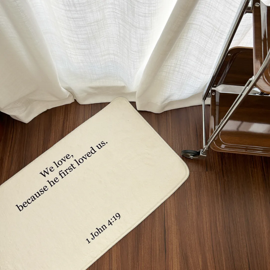 Cream rug with the words "We love, because he first loved us. 1 John 4:19" placed on a wooden floor by white curtains, bringing faith and simple happiness to any Christian home from Jincha God.