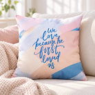 We Love Because He First Loved Us Christian cushion cover styled on a neutral sofa in a bright living room.