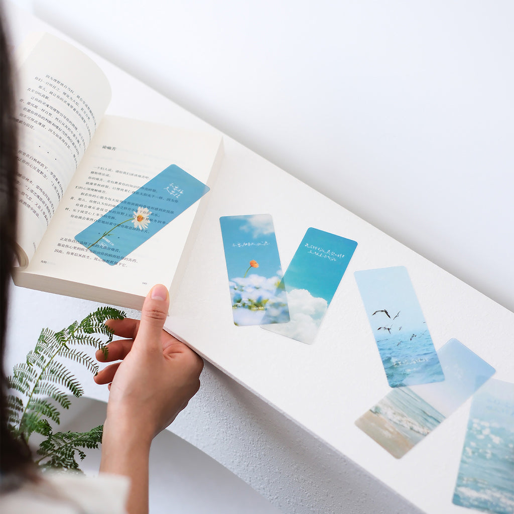 A woman reading a book with a Christian-themed bookmark featuring a daisy and Bible quote, alongside other bookmarks with ocean, sky, and flower designs, ideal for faith-inspired stationery.