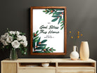 A beautifully styled wooden poster with the phrase "God Bless This Home and All Who Enter It," displayed on a wooden console table with a vase of white flowers and minimalist decor accents in the background.