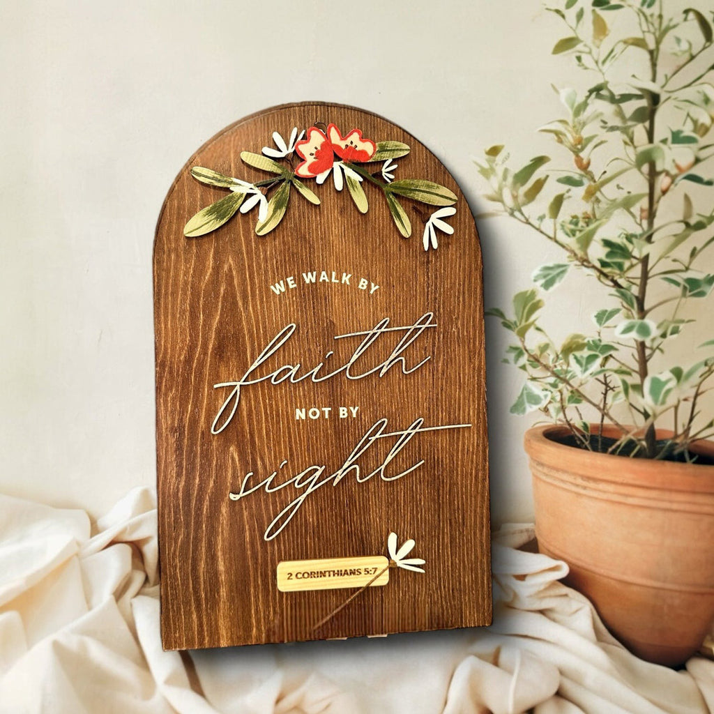 A wooden Christian wall art piece featuring the quote 'We walk by faith, not by sight' from 2 Corinthians 5:7. The arched design includes floral accents at the top, with engraved lettering in an elegant script. A potted plant sits next to the decor, complementing the natural wooden finish.