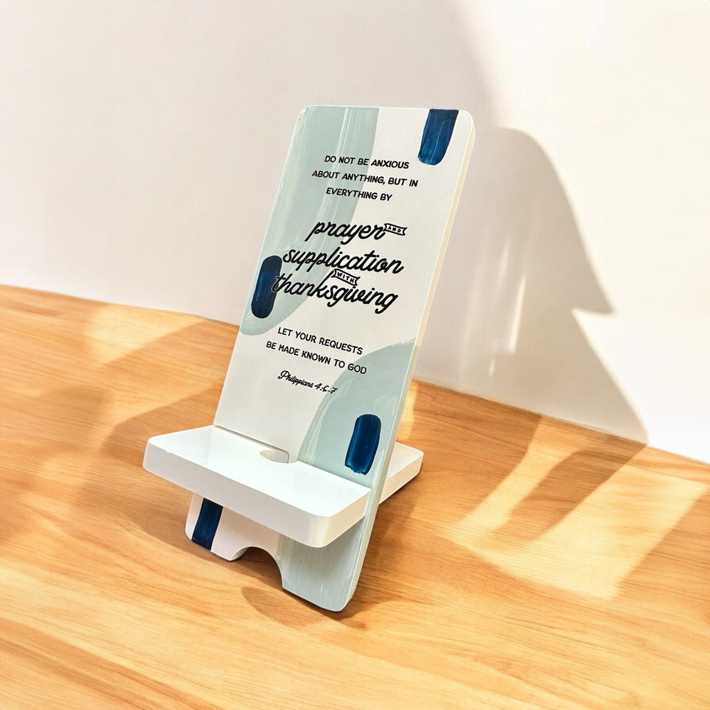 Wooden phone stand featuring the Bible verse Philippians 4:6 with text about prayer, supplication, and thanksgiving, placed on a wooden surface.