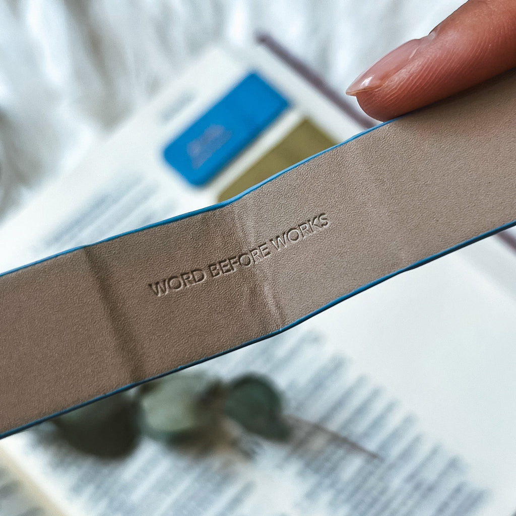 Close-up of a taupe-colored magnetic bookmark with 'Word Before Works' embossed, held over an open Bible with a blurred blue bookmark in the background.