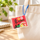 Xin Blessed Ramen acrylic keychain hanging from a cream canvas tote bag in bright natural light