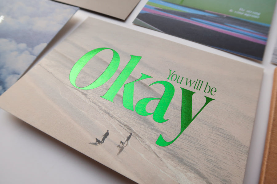 A close-up view of a collection of postcards, with a focus on the "You Will Be Okay" card featuring a serene beach background and bold green text, offering reassurance and encouragement.