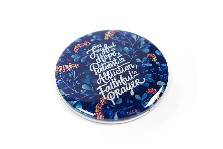 Close up of 5.5 cm diameter circular Acrylic fridge magnet with bible verse “Be joyful in Hope, patient in affliction, faithful in prayer” on foliage background.