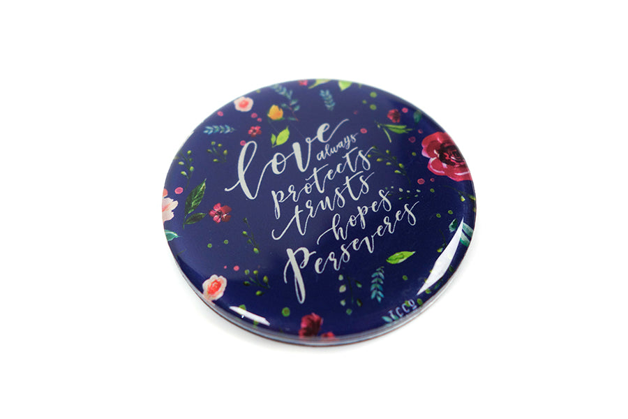 Close up of 5.5 cm diameter circular Acrylic fridge magnet with bible verse “Love always protects, trusts, hopes perseveres” on foliage background.