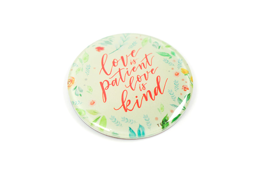 Close up of 5.5 cm diameter circular Acrylic fridge magnet with bible verse “Love is patient love is kind” on foliage background.